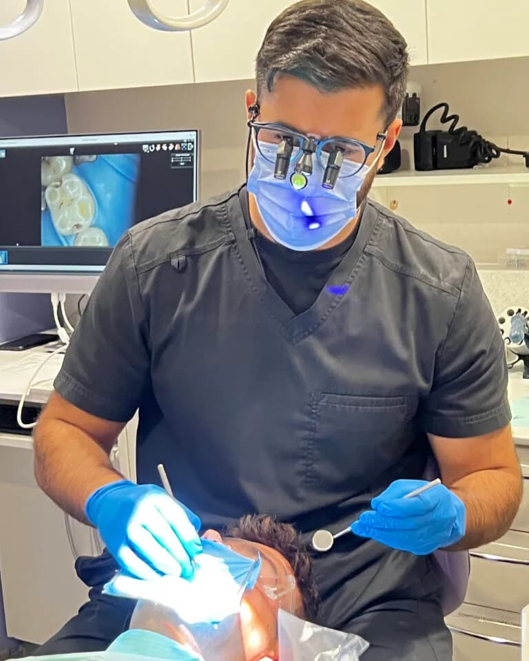 A dentist examining a patient's teeth using magnifying loupes, with the intra-oral camera image on screen
